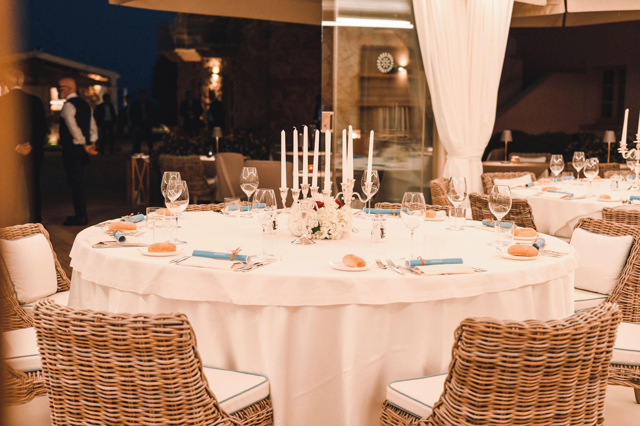 Une table de salle à manger ronde, recouverte d’une nappe blanche, entourée de chaises en osier et agrémentée de bougies.