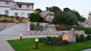 A landscaped garden with a stone pathway, a rock formation, and a building with multiple windows and balconies. - Hotel Borgo Smeraldo (Olbia)