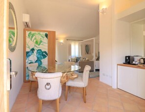 A dining area with a glass table and two chairs, a kitchen area with a coffee machine, and a living room with a sofa and a painting. - Hotel Borgo Smeraldo (Olbia)