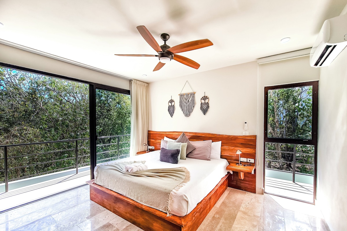 A spacious bedroom with a large bed, wooden headboard, and a ceiling fan. It features large windows offering a view of the outdoors.