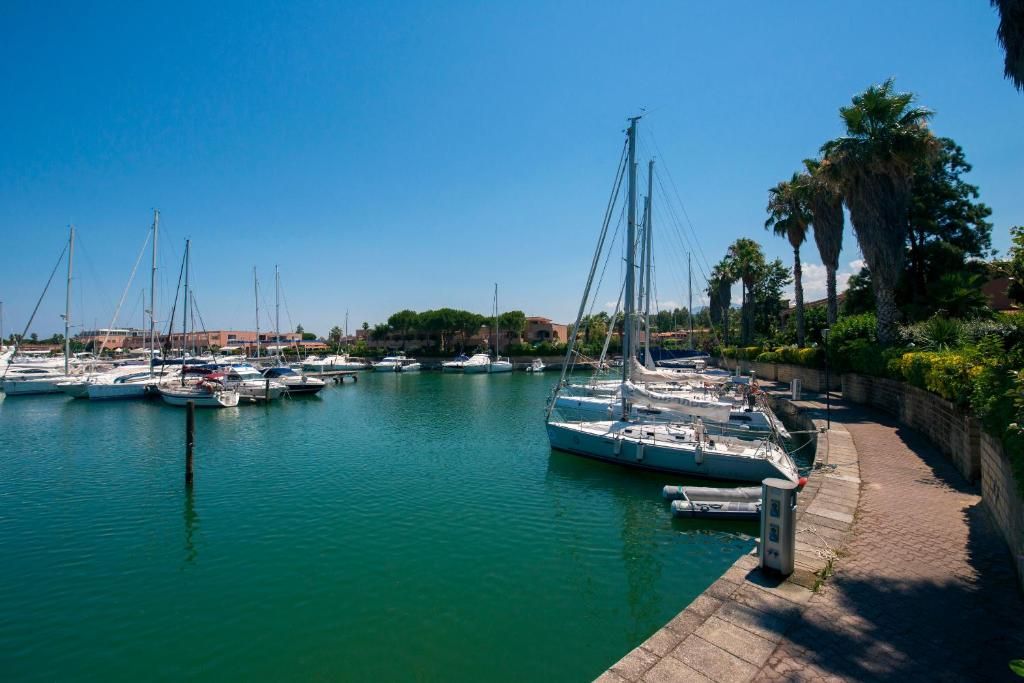 Ein Yachthafen mit mehreren Segelbooten, die an einer gepflasterten Uferpromenade liegen, umgeben von Grünflächen und Gebäuden im Hintergrund.