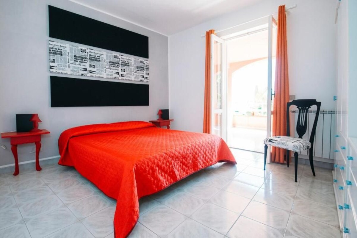 A bedroom with a large bed, a red blanket, a blackboard with text, a red side table, a chair, and a window with curtains.
