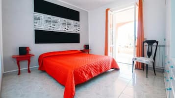 Une chambre avec un grand lit, une couverture rouge, un tableau noir avec du texte, une table de chevet rouge, une chaise et une fenêtre avec des rideaux.