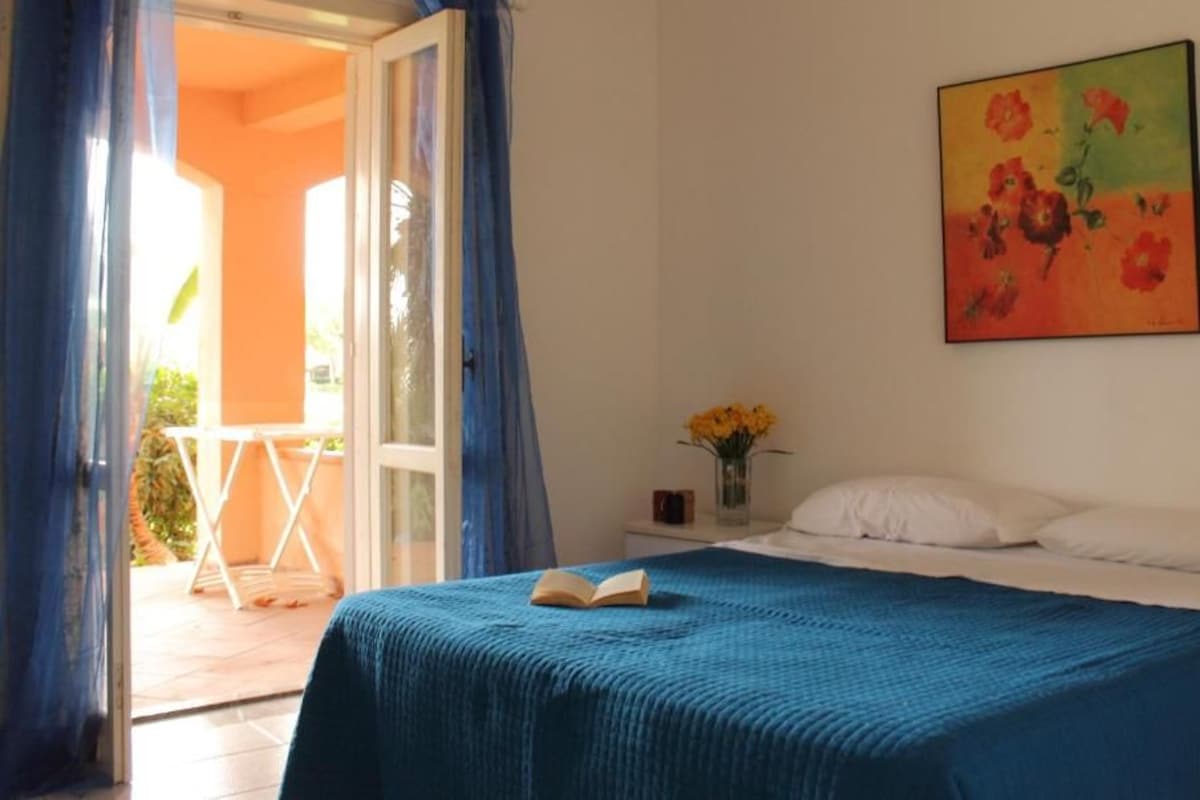A bedroom with a bed, a painting, a vase with flowers, and a view of the outside.
