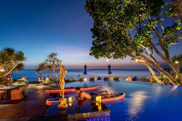 An infinity pool with a view of the ocean at dusk, surrounded by lounge chairs and a wooden deck.