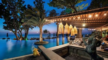 Zona de bar junto a la piscina con vista al océano, rodeada de árboles y luces colgantes.