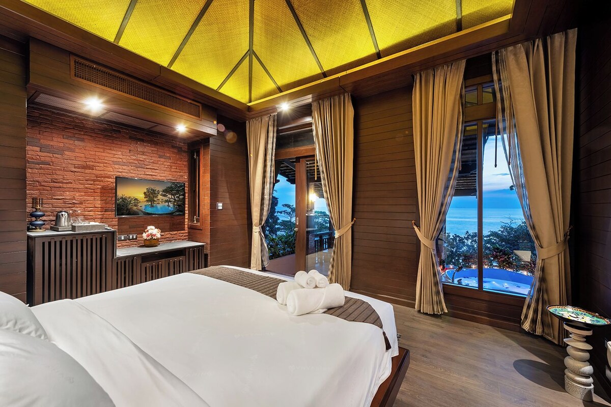 A hotel room with a large bed, a desk with a computer, a TV mounted on a brick wall, and a view of the ocean.