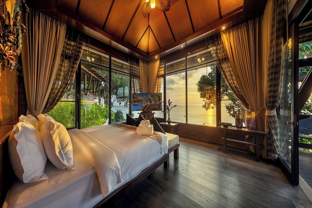 A spacious bedroom with a large bed, wooden flooring, and large windows offering a view of the outdoors.