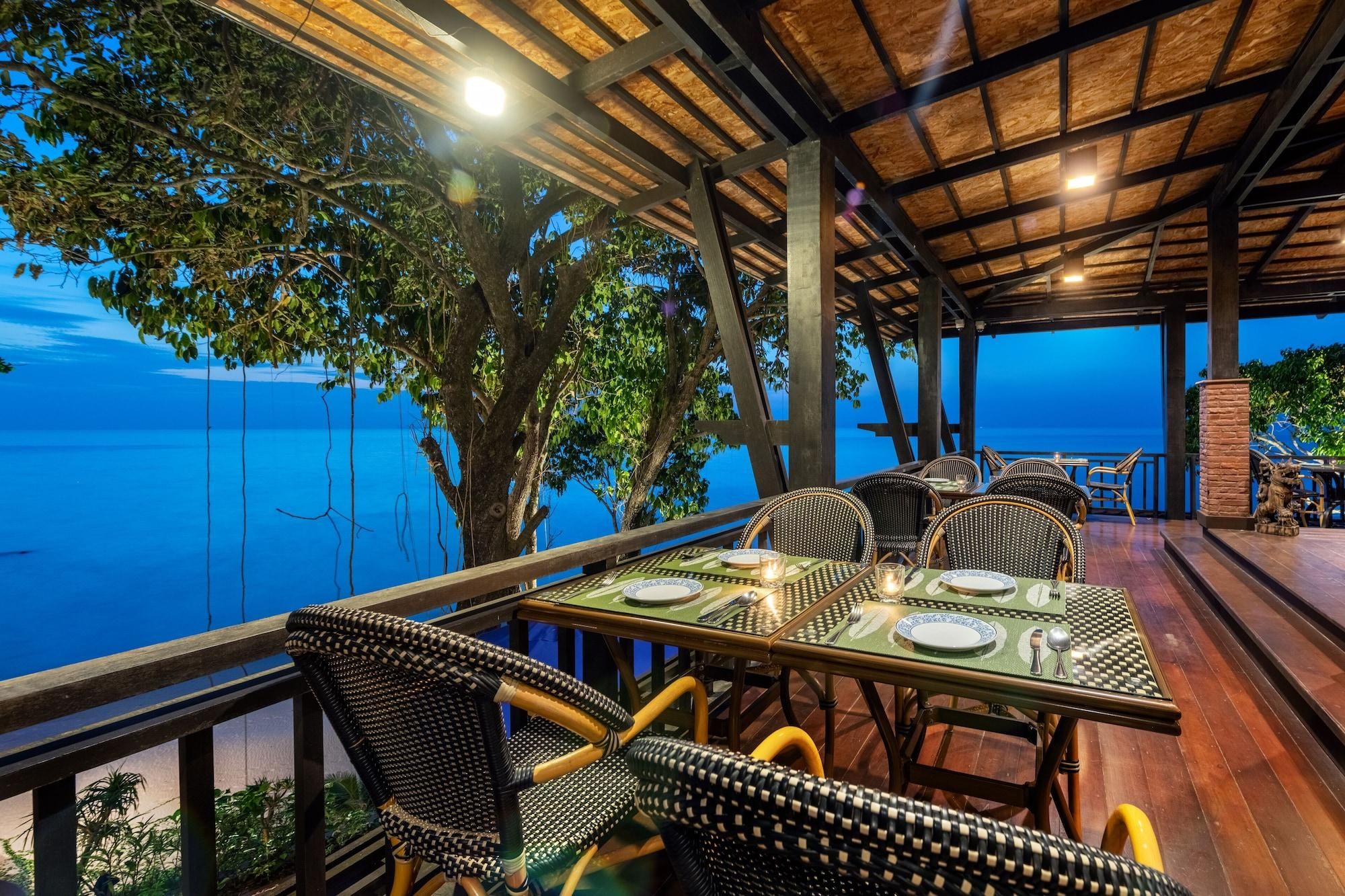 Ein Essbereich im Freien mit Blick auf das Meer, ausgestattet mit Korbstühlen und Tischen, gedeckt für ein Essen.