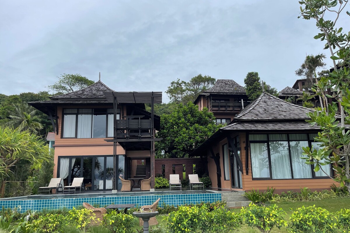 A two-story house with a balcony, a swimming pool, and lush greenery.