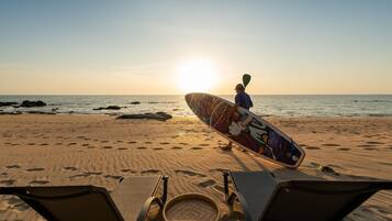 Escena de playa con una persona cargando una tabla de surf colorida, dos tumbonas y una cesta tejida sobre la arena.