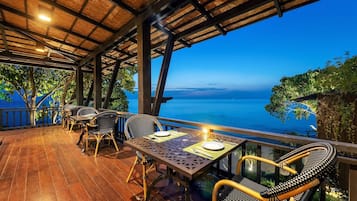 Un espacio de comedor al aire libre con piso de madera, mesas metálicas y sillas de mimbre, con vista a un cuerpo de agua.