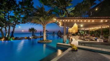 Zona de bar junto a la piscina con vista al océano, rodeada de palmeras y un área de estar cubierta con luces colgantes.