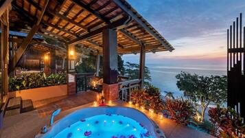 Un jacuzzi con nenúfares, una pérgola de madera y vista al océano al atardecer.