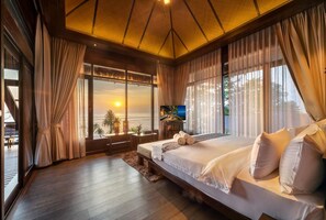 Een slaapkamer met een groot bed, een televisie en uitzicht op de ondergaande zon boven de oceaan.