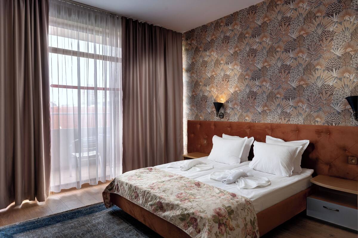 Une chambre d’hôtel avec un grand lit, une tête de lit en bois, un mur à motifs, une fenêtre avec des rideaux légers et une table de chevet avec une lampe.