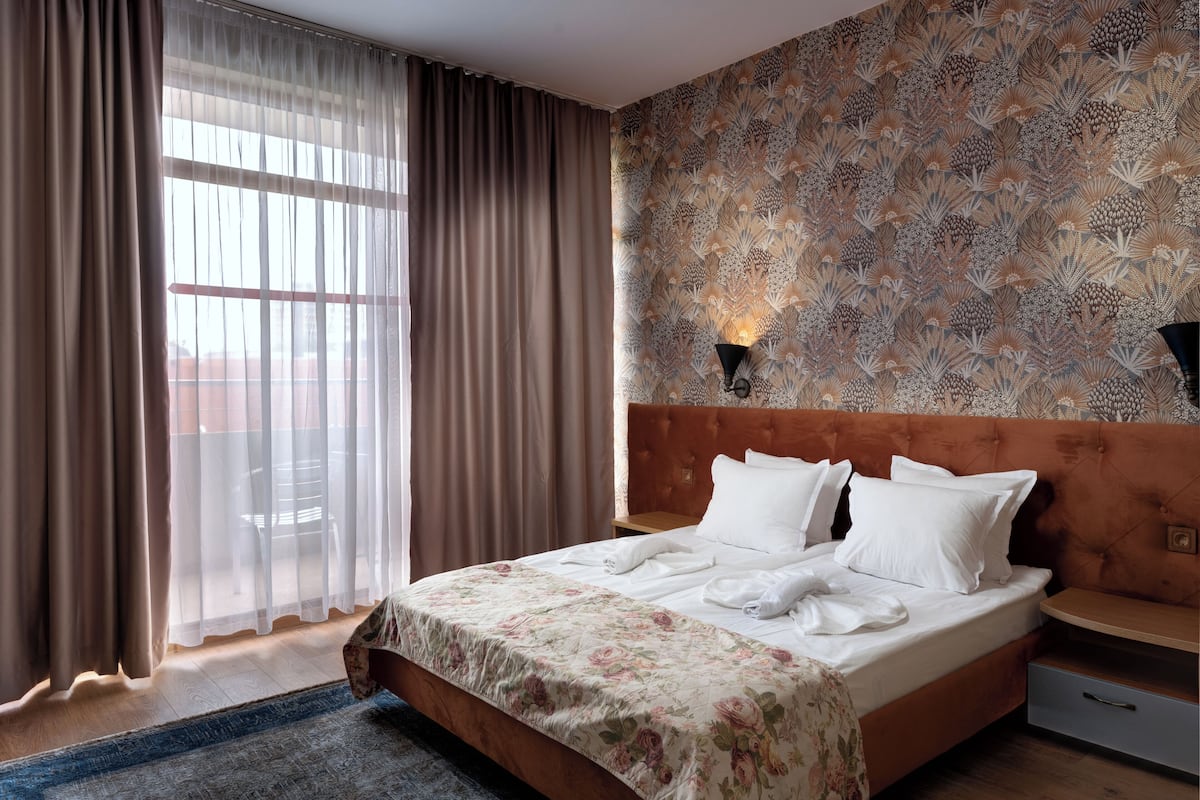 Una camera d'albergo con un letto grande, una testiera in legno, una parete decorata con un motivo, una finestra con tende leggere e un comodino con una lampada.