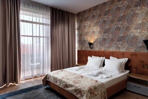 A hotel room with a large bed, a wooden headboard, a patterned wall, a window with sheer curtains, and a nightstand with a lamp. - Avenue (Nessebar)