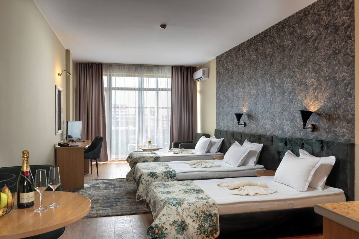 A hotel room with three beds, a desk, a television, and a view of the city.