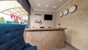 A reception area with a blue chair, a reception desk, three wall clocks, a flat-screen TV, and an air conditioning unit. - Avenue (Nessebar)
