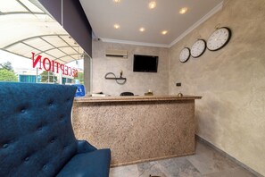 A reception area with a blue chair, a reception desk, three wall clocks, a flat-screen TV, and an air conditioning unit. - Avenue (Nessebar)