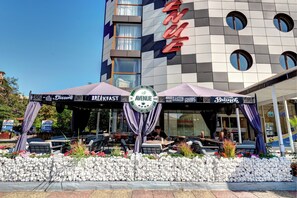 A hotel with a modern facade, outdoor seating area, and a sign indicating breakfast service. - Avenue (Nessebar)