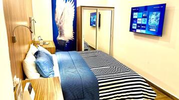 A hotel room with a bed, a desk, a mirror, and a wall-mounted TV displaying streaming services.