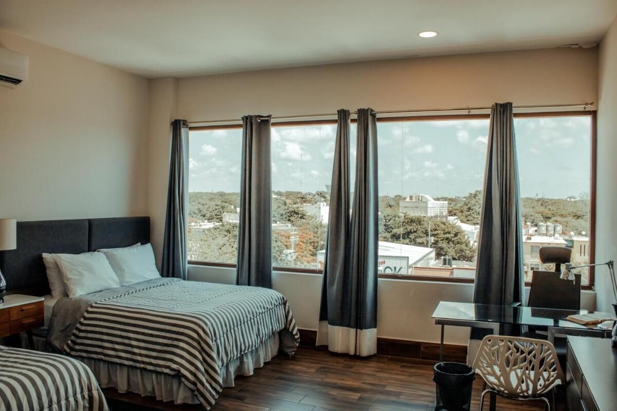 A hotel room with a large window, a bed, a desk, and a chair.