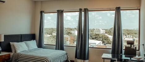 A hotel room with a large window, a bed, a desk, and a chair.