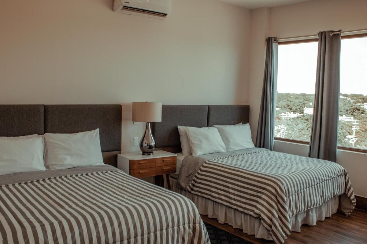 A hotel room with two beds, a nightstand, a lamp, and a window with curtains.
