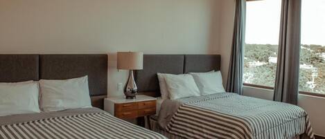 Habitación de hotel con dos camas, una mesita de noche, una lámpara y un ventanal con cortinas.