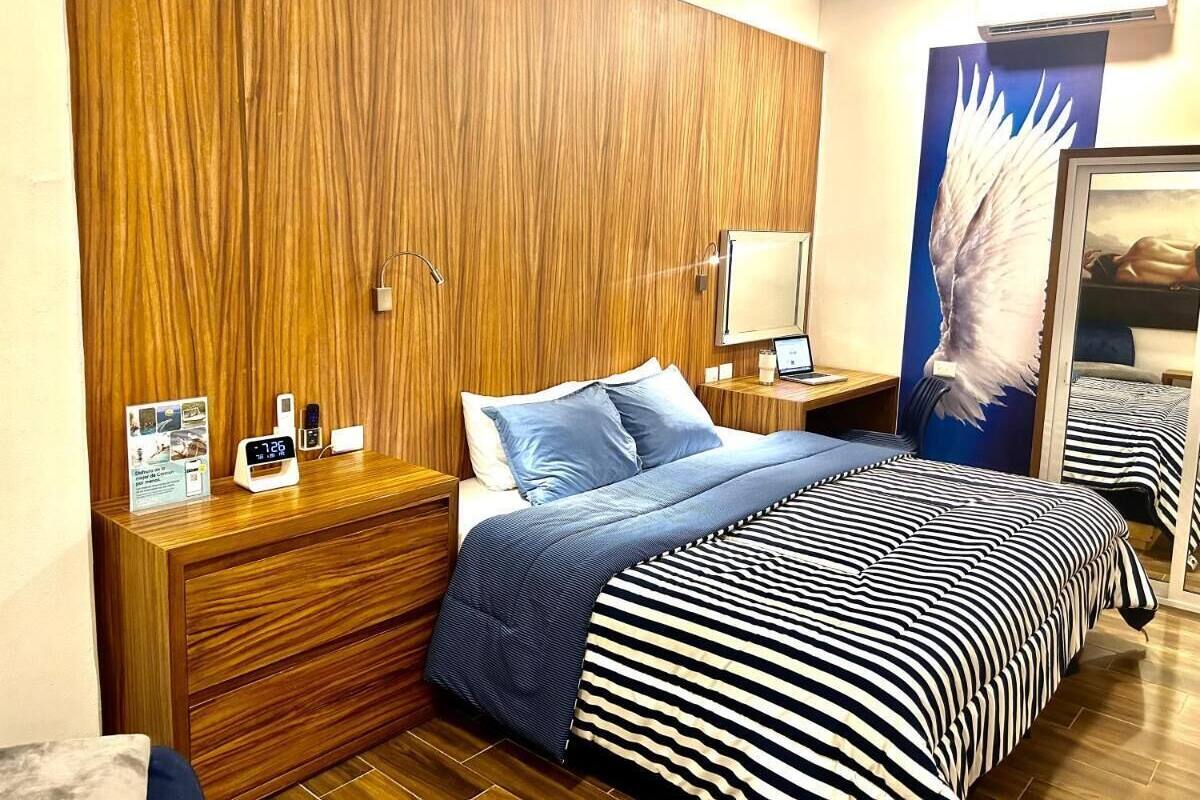 Une chambre d’hôtel avec une tête de lit en bois, un lit avec une literie à rayures, une table de chevet avec une horloge et un cadre photo, et un miroir au mur.
