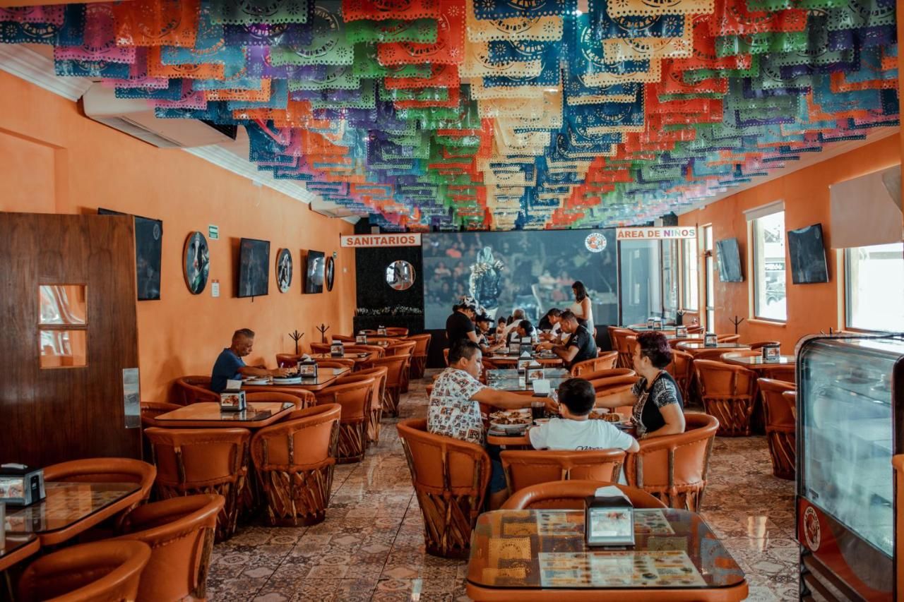 A restaurant with orange walls, wooden tables, and chairs. There are signs indicating 'SANITARIOS' and 'AREA DE NIÑOS'.