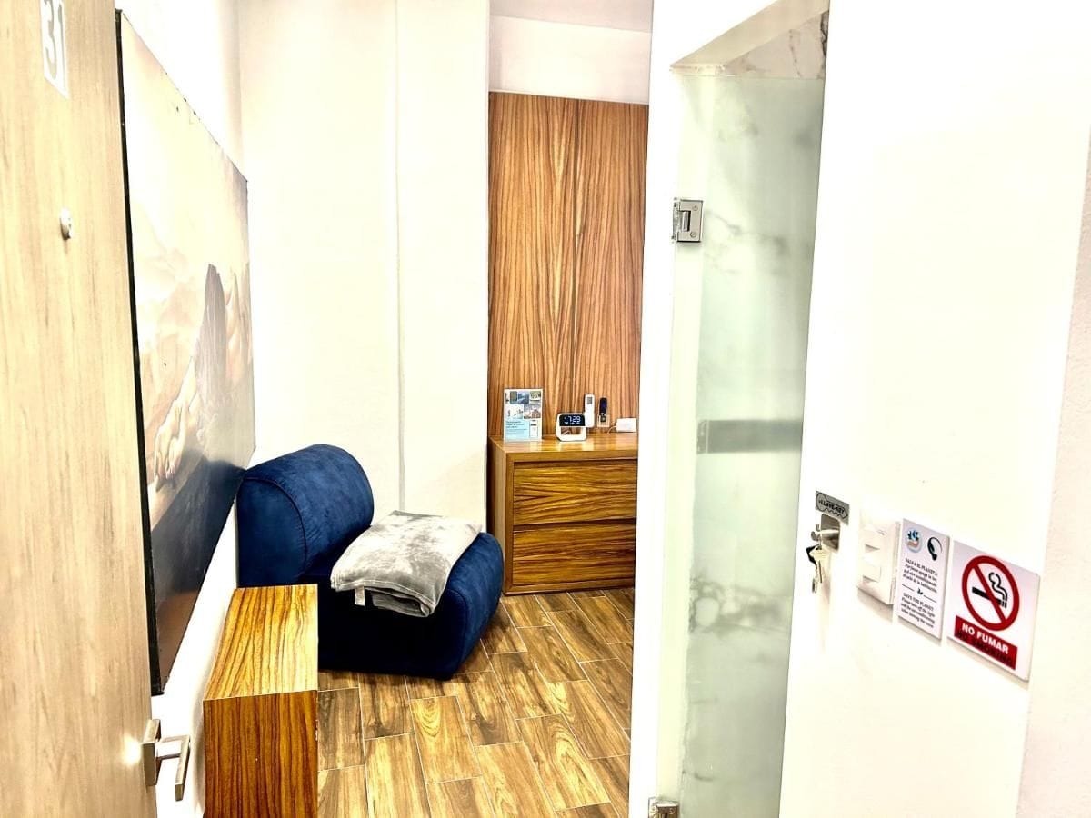 A hotel room with a wooden floor, a blue sofa, a small table with a blanket, and a wooden cabinet with a clock and a phone.