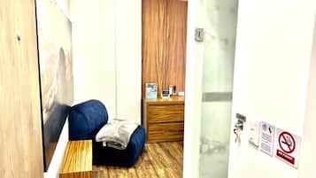 A hotel room with a wooden floor, a blue sofa, a small table with a blanket, and a wooden cabinet with a clock and a phone.