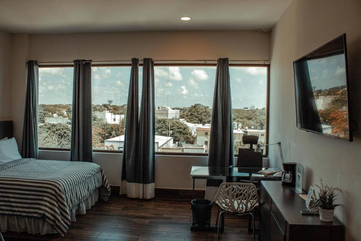 Une chambre d’hôtel équipée d’un lit, d’un bureau, d’une chaise et de grandes fenêtres donnant sur la ville.
