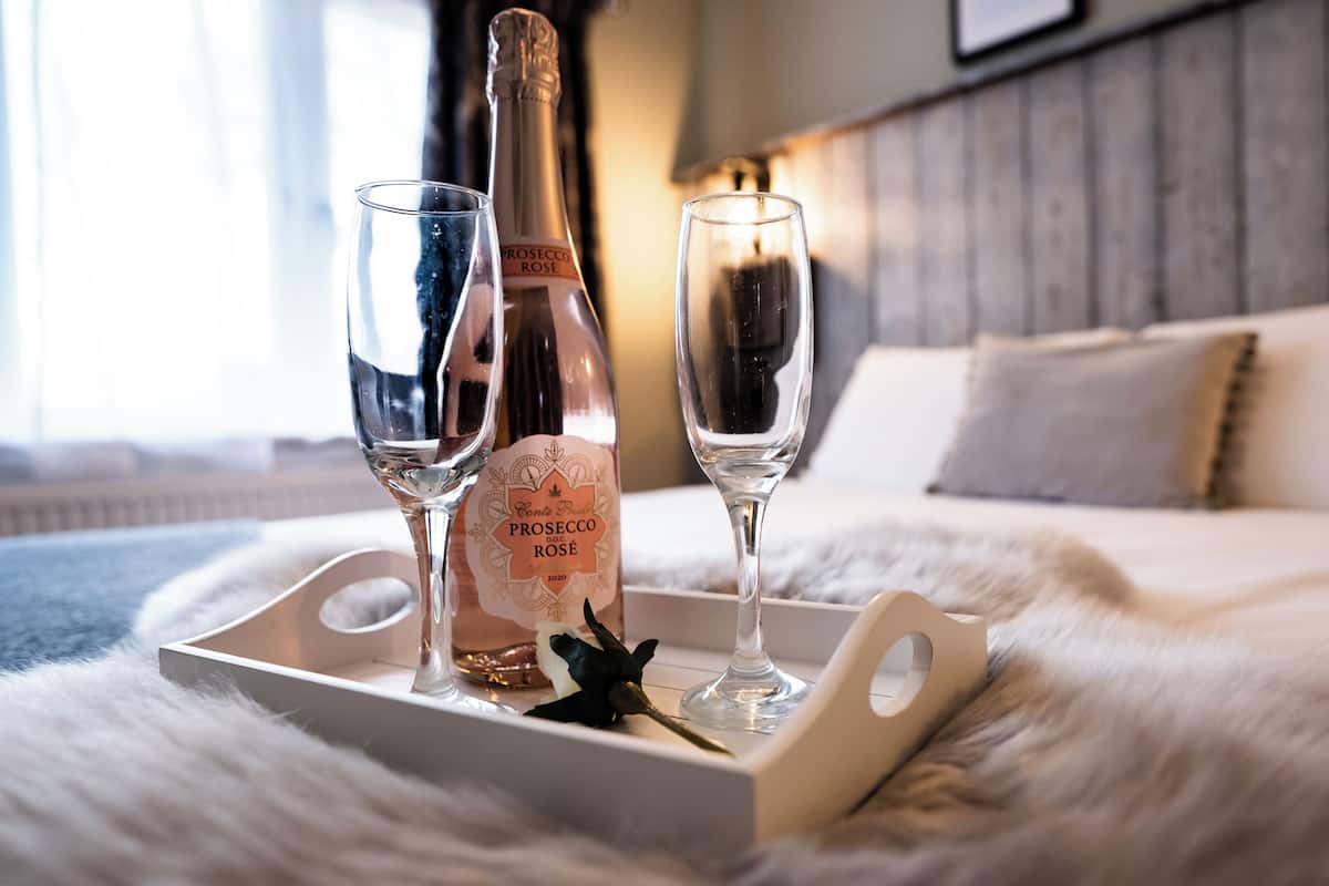 A bottle of Prosecco DOC G Rose on a tray with two champagne flutes on a fluffy white bedspread.