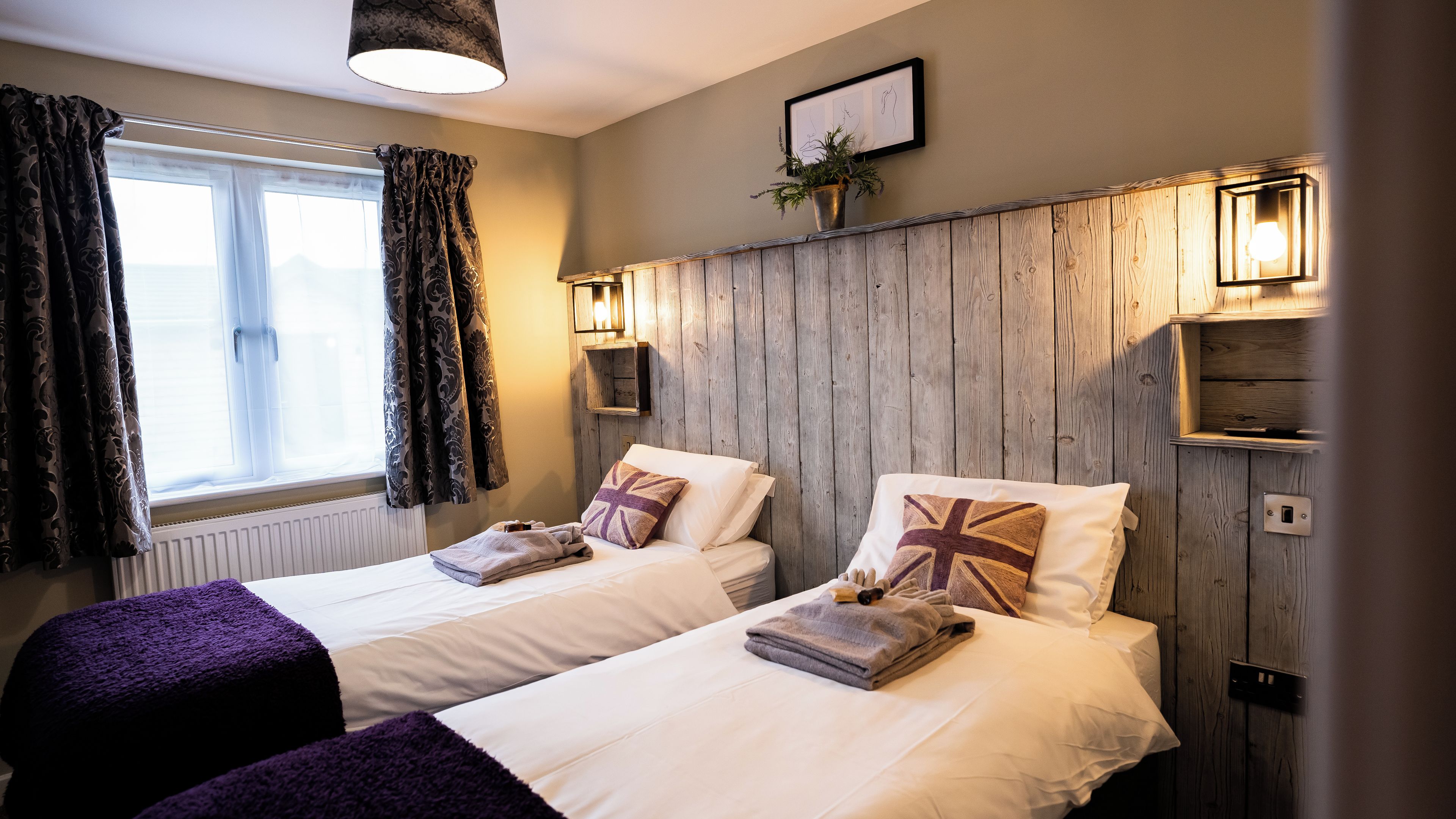 A room with two beds, each with a Union Jack pillow, a window with curtains, and wall-mounted lights.