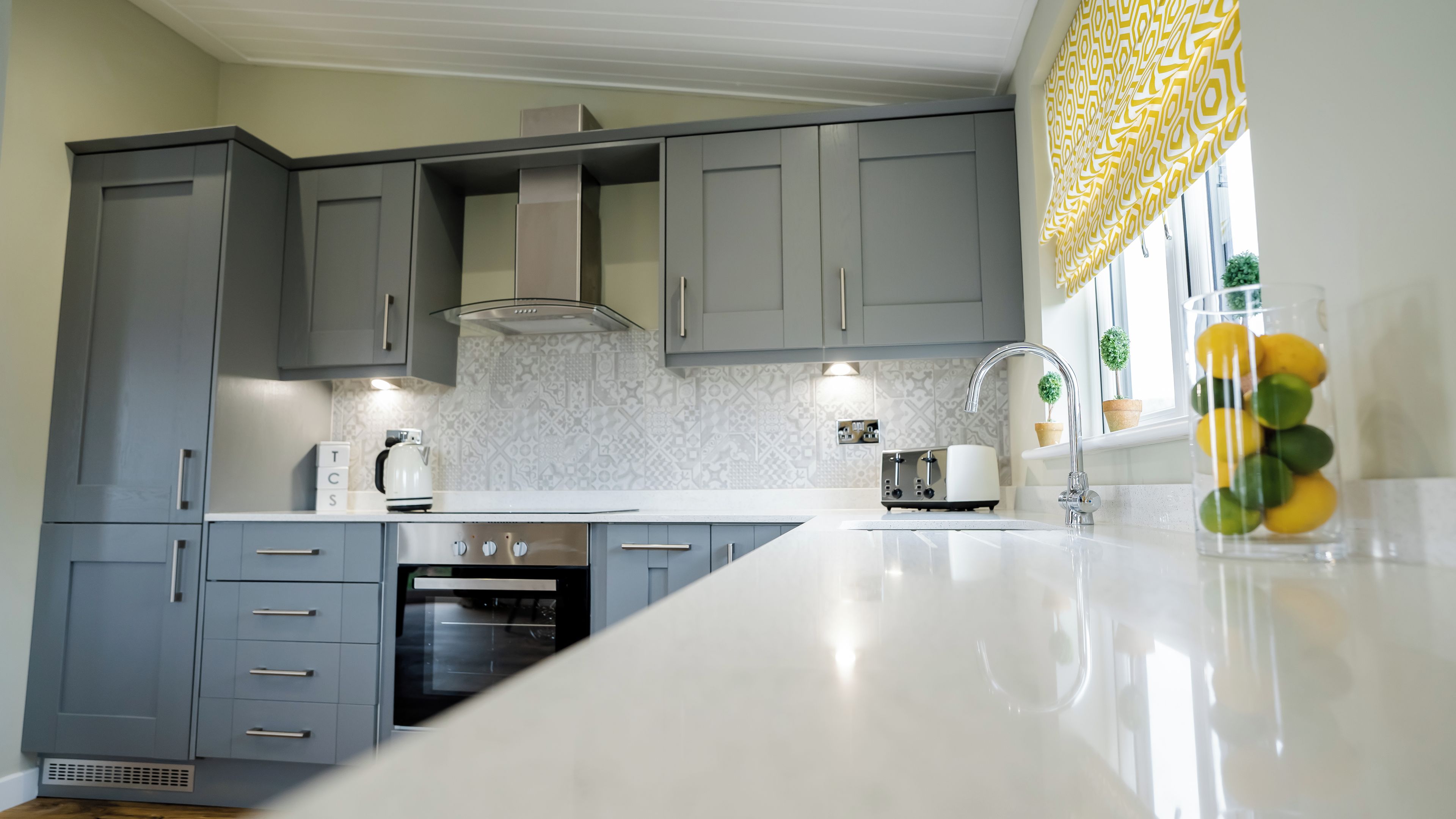 Una cocina moderna con gabinetes grises, encimera blanca, horno empotrado y una ventana con cortinas de estampado amarillo.