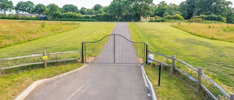 Un chemin pavé mène à une entrée fermée par un portail, qui est elle-même entourée de champs verdoyants et d’arbres.