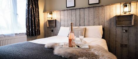 A hotel room with a wooden headboard, a bed with white linens, a fur throw, a tray with a bottle and glasses, and framed pictures on the wall.