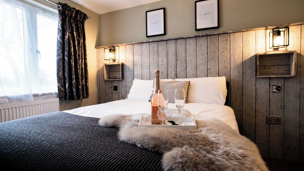 A hotel room with a wooden headboard, a bed with white linens, a fur throw, a tray with a bottle and glasses, and framed pictures on the wall. - Chestnut Meadow Country Park (Bexhill-on-Sea)