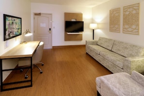 A hotel room with a wooden desk, a sofa, a television mounted on the wall, and a door with a sign.