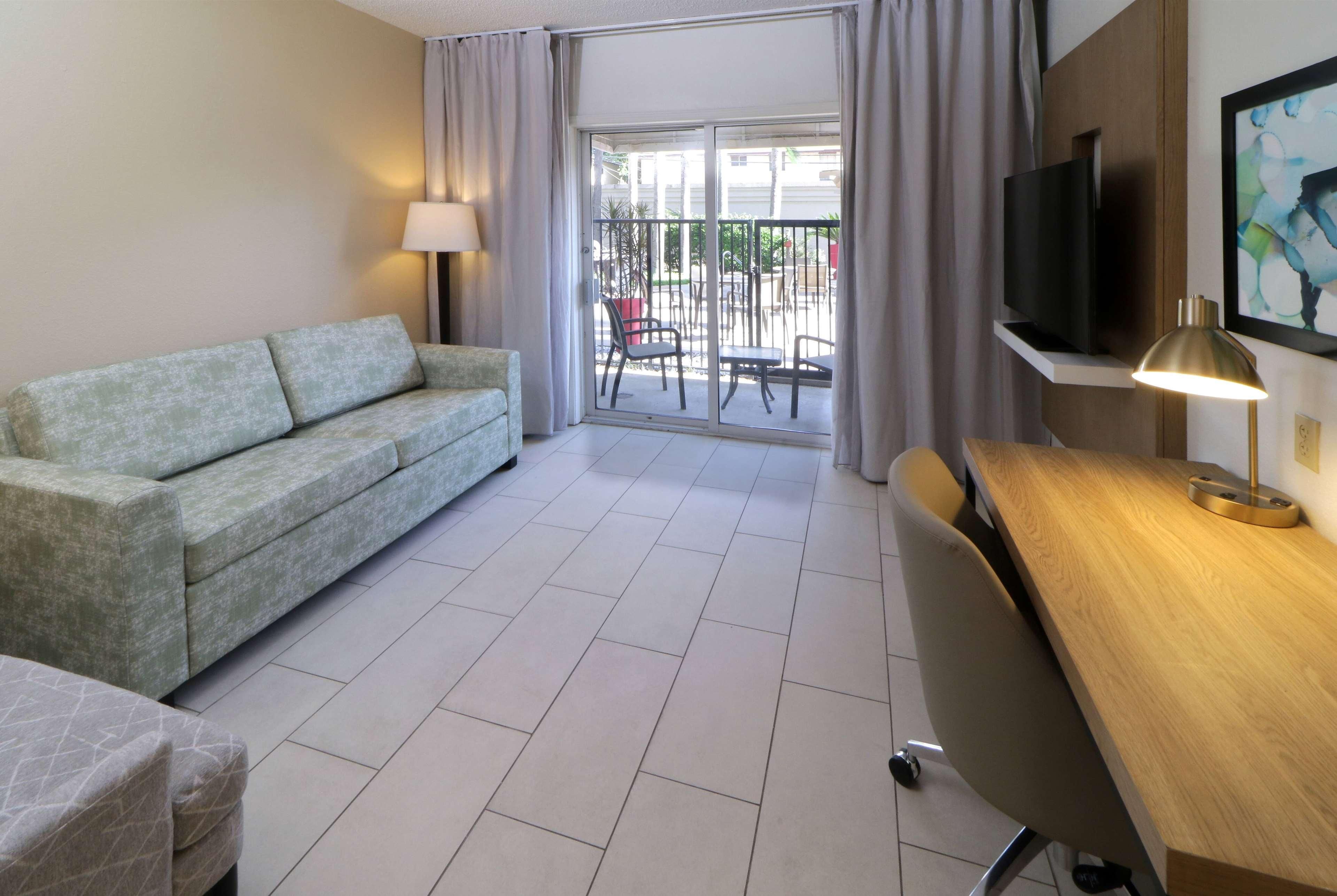 A hotel room with a sofa, a desk with a lamp, a TV, and a view of the outside balcony.