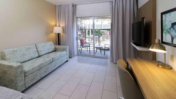 A hotel room with a sofa, a desk with a lamp, a TV, and a view of the outside balcony.