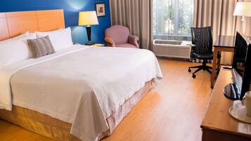 A hotel room with a large bed, a desk with a computer, a chair, and a window with curtains.