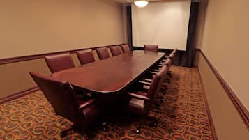 Un salón de conferencias con una larga mesa y varias sillas, piso alfombrado y una pared lisa con una lámpara.