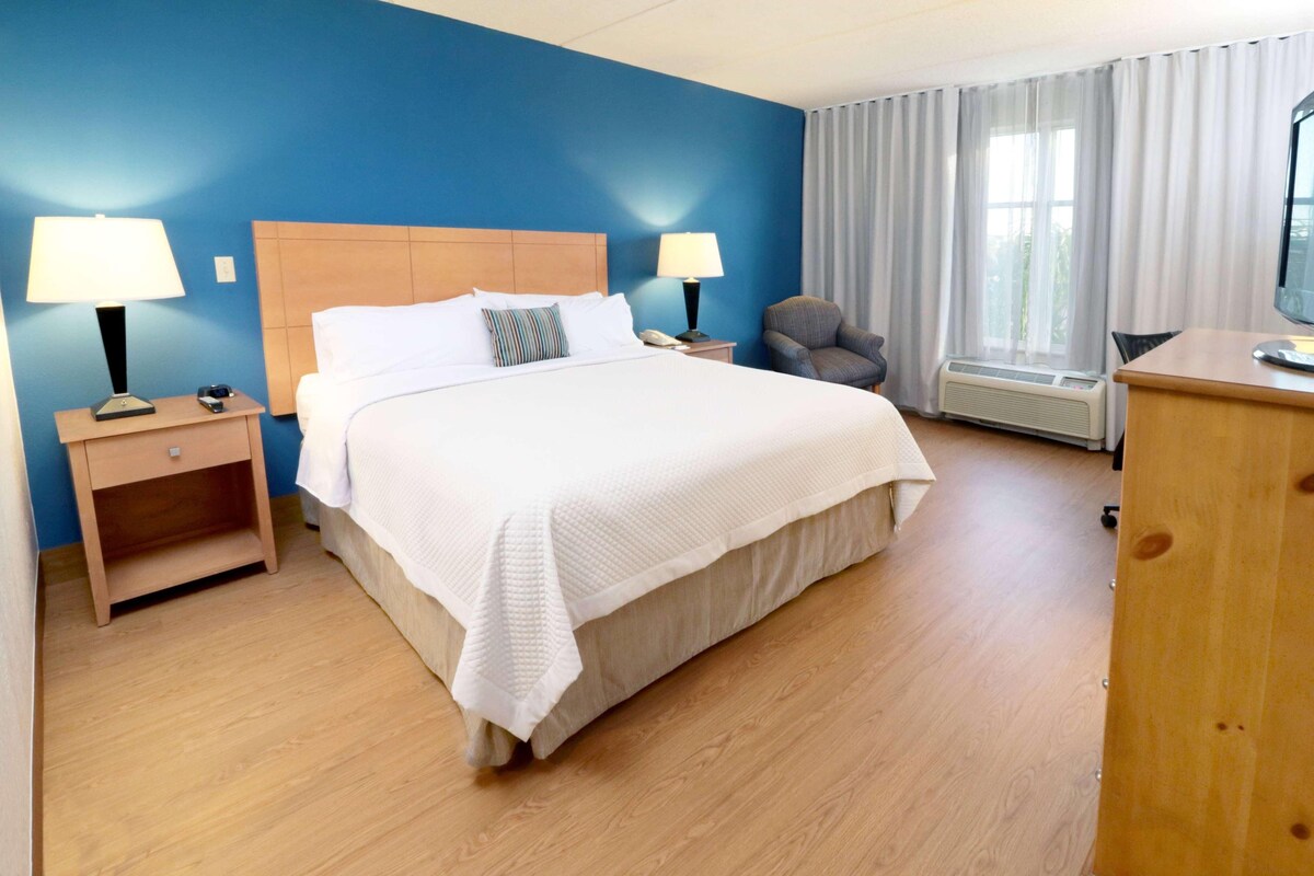 Ein Hotelzimmer mit einem großen Bett, zwei Nachttischen mit Lampen, einem Sessel, einem Schreibtisch und einem Fenster mit Vorhängen.