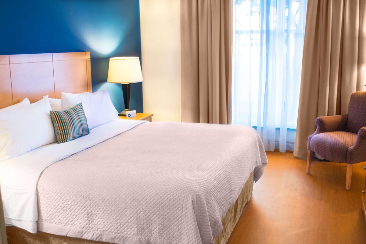 Ein ordentlich gemachtes Bett mit weißen Leinen und einem gestreiften Kopfkissen, eine Nachttischlampe, ein Sessel und ein Blick ins Freie durch transparente Vorhänge.