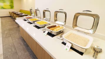 A buffet line with various food items in metal chafing dishes, a seating area with chairs, and a green cushioned bench.